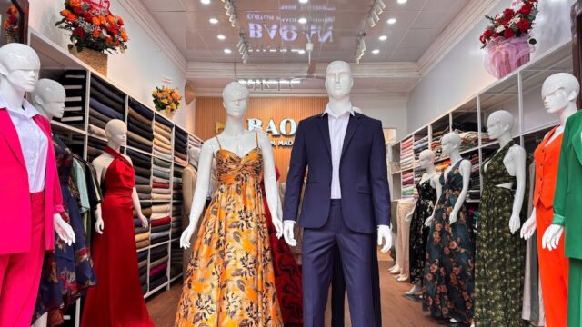 Mannequins in formal dresses and suits at Bao An 2 Tailor, a brightly lit fabric store adorned with elegant flower arrangements.