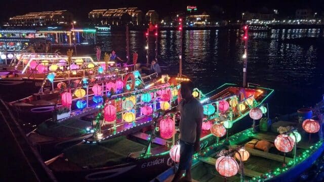 Colorful boats from Bao Long Boat Tour, decorated with glowing lanterns, float on a river at night in a vibrant riverside town.