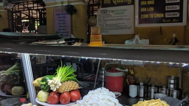 Plates of Vietnamese noodles, shrimp, and fresh vegetables displayed at a market food stall with signs behind.