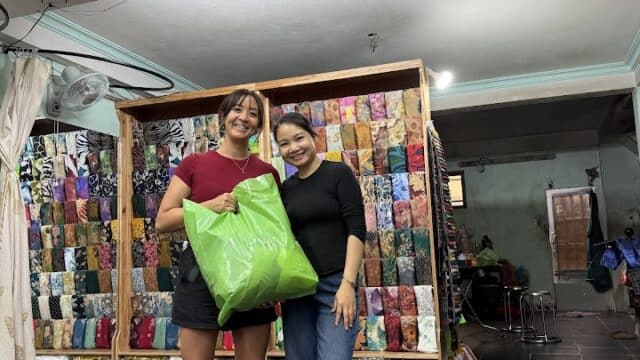 Two women smiling in front of Be Silk Tailor shop in Hoi An, one with a large green bag, fabrics displayed behind them.