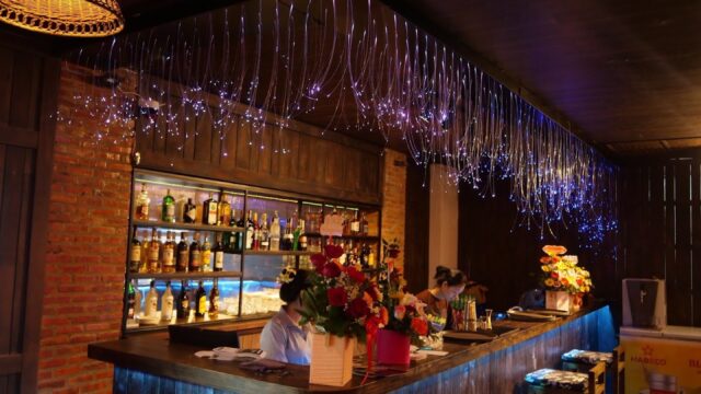 A cozy Hoi An bar with glowing string lights, flowers on the counter, and bottles displayed on wooden shelves.