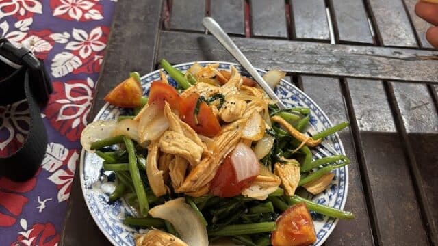 A plate of stir-fried chicken with vegetables, inspired by Hoi An, sits on a wooden table beside chopsticks and a patterned tablecloth.