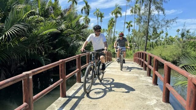 Two people ride bicycles across a small bridge in Hoi An, surrounded by lush green palm trees on a sunny day.