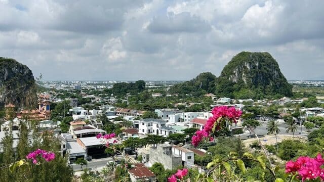 Pink flowers in the foreground overlook a city and green hills, capturing the charm of Vietnam Travel beneath a bright, partly cloudy sky.