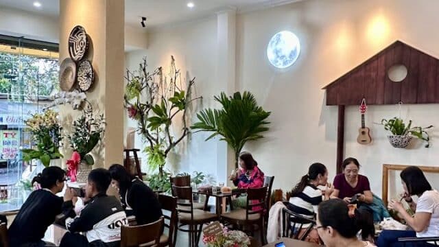 People sitting at tables in a cozy Hoi An café, chatting, eating, and using their phones, surrounded by plants and wall decor.