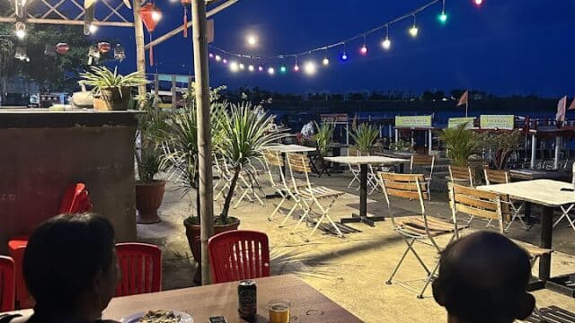 Two people sit at an outdoor table at dusk under string lights at Bi's Dock restaurant, with empty chairs and tables nearby.