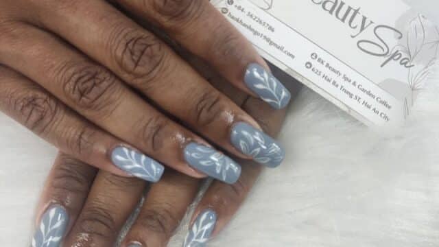Hands with long, blue nails featuring white leaf designs, holding a BK Beauty Spa business card at the beauty spa.