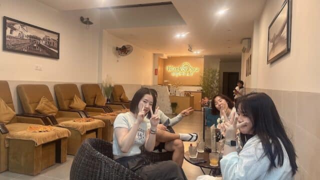 Four people relax and pose for a photo in a cozy spa lounge with warm lighting and empty massage chairs.