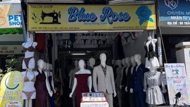 A tailor shop named Blue Rose in Hoi An with mannequins in suits and dresses displayed outside on a sunny day.