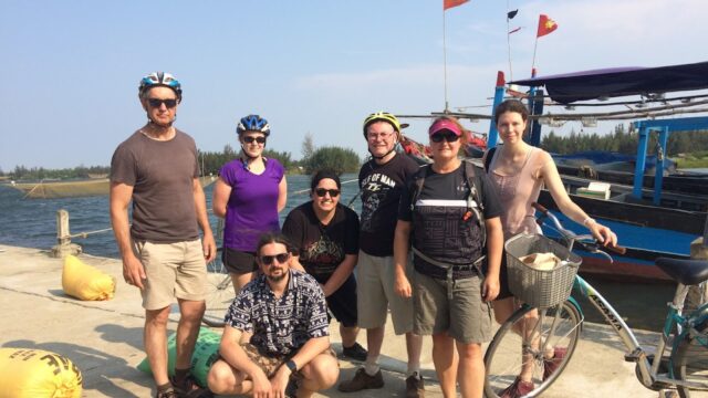 Six people with bikes pose on a sunny dock near water in Hoi An, with flags and boats in the background.