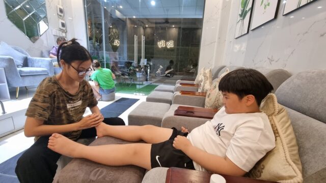A woman massages a boy's legs as he relaxes on a chair in a modern, well-lit spa, enjoying ultimate comfort and care.