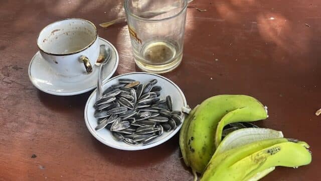 A coffee cup, glass, sunflower seeds, and two bananas on a wooden table outdoors.