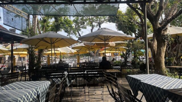 Outdoor cafe Cà Phê Vọng Phố with empty tables, checkered tablecloths, yellow umbrellas; one person sits alone.