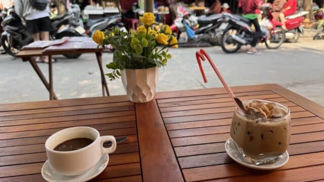 Two drinks and a vase of yellow flowers on a wooden table at a street-side café with scooters passing by.