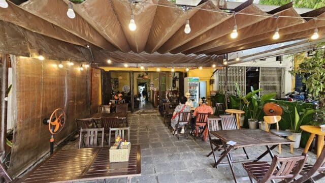 Outdoor café with wooden tables, string lights, potted plants, and two people sitting near the entrance.