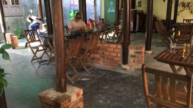 A man sits alone at a wooden table in a shaded, open-air Hoi An café with empty chairs and scattered bricks.
