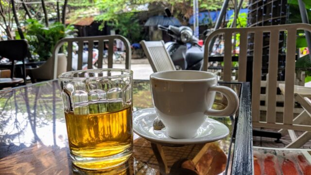 A glass of tea and a cup of coffee sit on a glass table at an outdoor café with chairs and greenery.