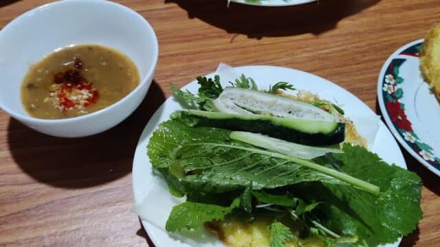 Plate with fresh herbs, cucumber, and pancake beside a bowl of dipping sauce, evoking the flavors of Hoi An on a wooden table.