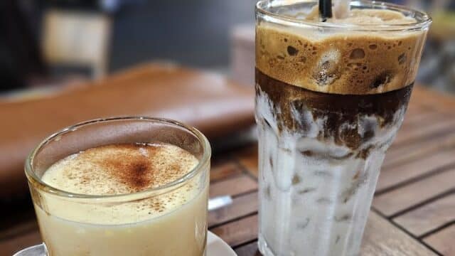Two Vietnamese coffee drinks on a wooden table in Hoi An, one hot with foam and one iced with layers and straws.