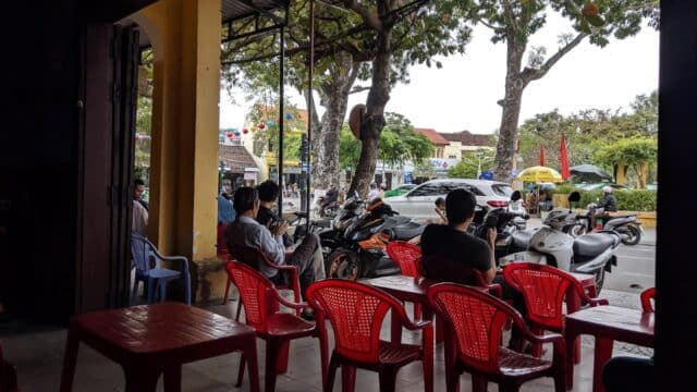 People sit on red plastic chairs at an outdoor cafe in Hoi An, with parked motorbikes and trees in the background.