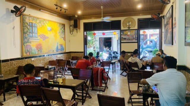 People sit on wooden chairs in a cozy Hoi An café with wall art, facing the street through large front windows.
