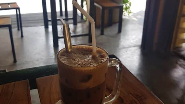 Iced coffee in a glass mug with a spoon and straw, on a wooden table at a café with an open street view.