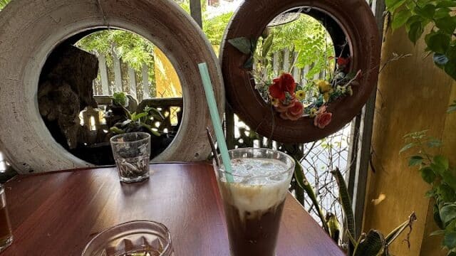 Iced coffee and tea on a wooden table in Hoi An, beside a garden with hanging plants and tire planters.