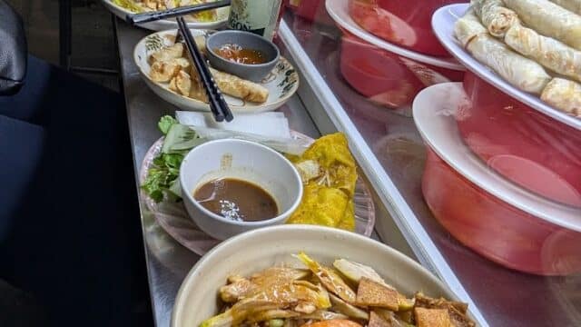 Vietnamese street food dishes from Hoi An with grilled meats, shrimp, and vegetables on a metal counter beside drinks and condiments.