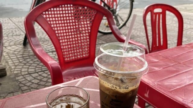 Glass of tea and iced coffee at Cafe Muối Min on a red table outdoors, with red chairs and a bicycle in the background.