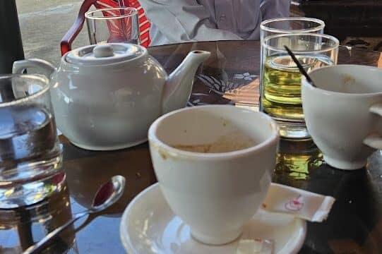 An elderly man sits at an outdoor cafe table in Hoi An, with tea, water, and coffee cups in the morning sunlight.
