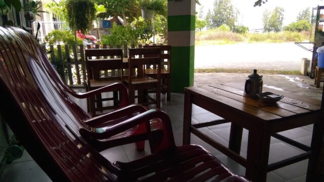 Empty wooden chairs and a table with a teapot on a shaded porch in Hoi An, overlooking a sunny outdoor scene.