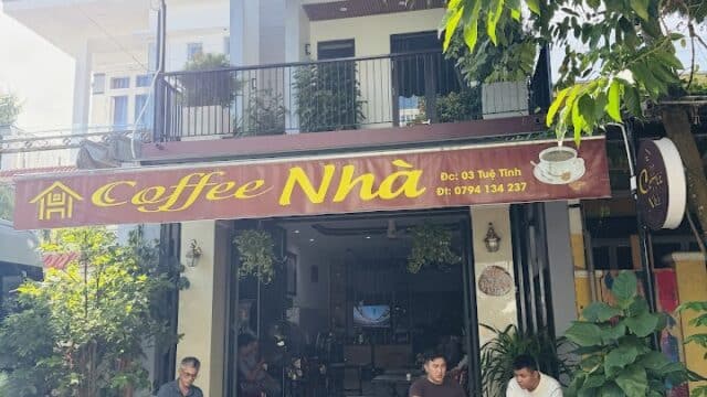People sitting outside Coffee Nhà café in Hoi An under trees, with scooters parked in front on a sunny day.