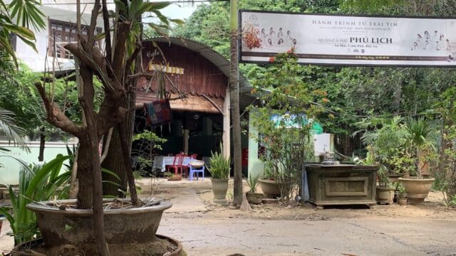 A rustic house with palm trees, potted plants, and a large Cafe Phú Lịch sign in Vietnamese at the entrance.
