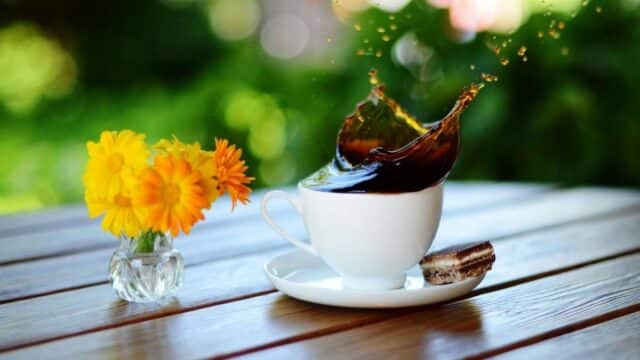 Coffee splashing out of a white cup at Cafe Que Huong on a wooden table, with yellow flowers and a cookie nearby.