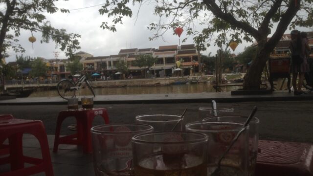 Glasses on a red table at an outdoor café by the river in Hoi An, with trees and lanterns glowing in the background.