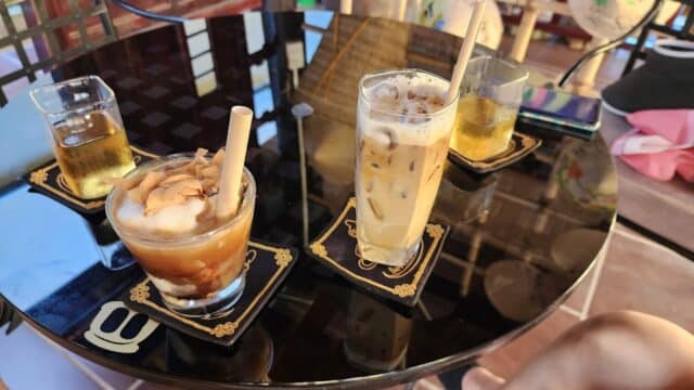 Four iced drinks on a reflective black table at Cafe Seven Dwarfs, with two people sitting nearby on a tiled floor.