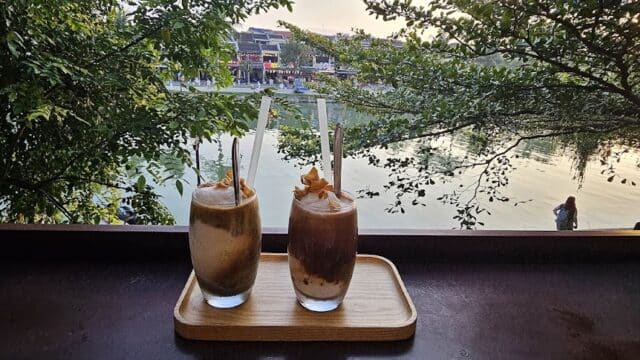 Two iced drinks with straws on a wooden tray, overlooking a riverside scene with trees and people at sunset.