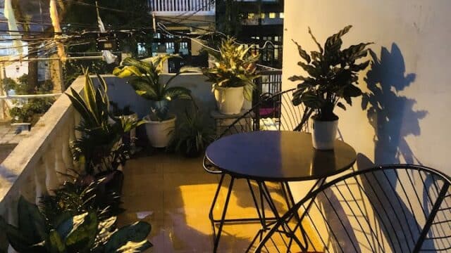 A cozy Hoi An balcony at dusk with plants, a round table, and two chairs with orange cushions.
