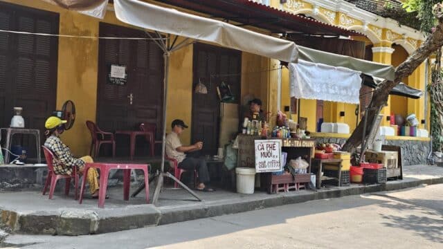 Street coffee stand with two men sitting and a woman nearby, set against a yellow building on a sunny day.
