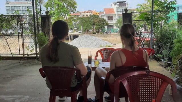 Two people sit on red chairs at Cafe Vong Vuon Ngoc, enjoying drinks and a riverside view beneath a cozy roof.
