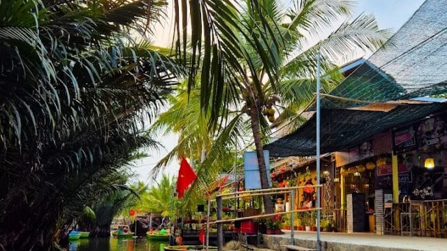Round basket boats docked by a canal in Cam Thanh Coconut Village, lined with palm trees, shops, and a red flag under a partly cloudy sky.