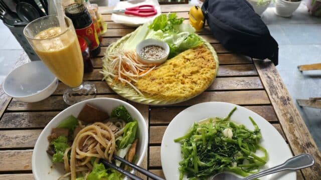 Vietnamese meal on a wooden table in Hoi An with pancakes, noodles, greens, dipping sauce, and a glass of orange drink.