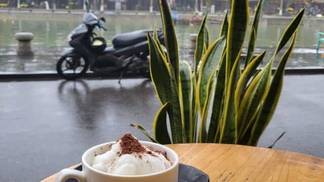 A cup of cappuccino on a wooden table by Hoi An’s rainy riverside street, with plants and a parked motorbike nearby.