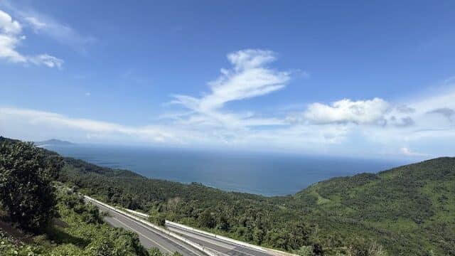 A winding mountain road in Central Vietnam with a motorbike, lush greenery, and sea views under a bright blue sky.