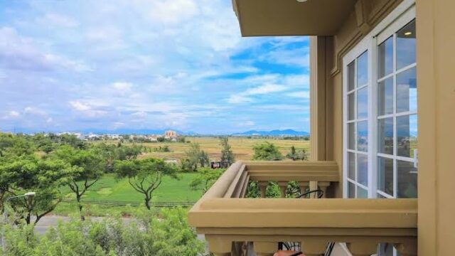 A balcony at CGP Luxury Hotel with chairs overlooks green fields and trees under a bright blue sky with scattered clouds.