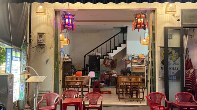 A small, open-front cafe in Hoi An with red plastic chairs, wooden tables, and hanging lanterns at dusk.
