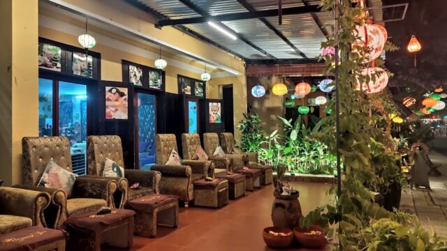 Row of plush chairs with pillows, colorful lanterns, and plants in a cozy, warmly-lit indoor-outdoor lounge.