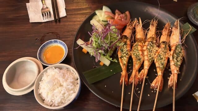 Grilled shrimp skewers with rice, dipping sauce, and fresh vegetable salad at Cho Que Riverside Restaurant on a dark wooden table.