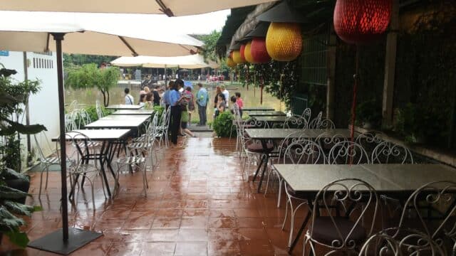 Outdoor café with empty tables, wet tiled floor, umbrellas, red lanterns, and people gathered in the background.