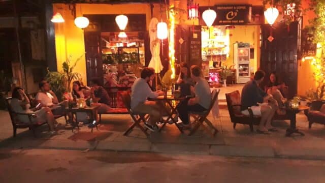 People sitting at outdoor tables, eating and chatting at a warmly lit restaurant and coffee bar in the evening.